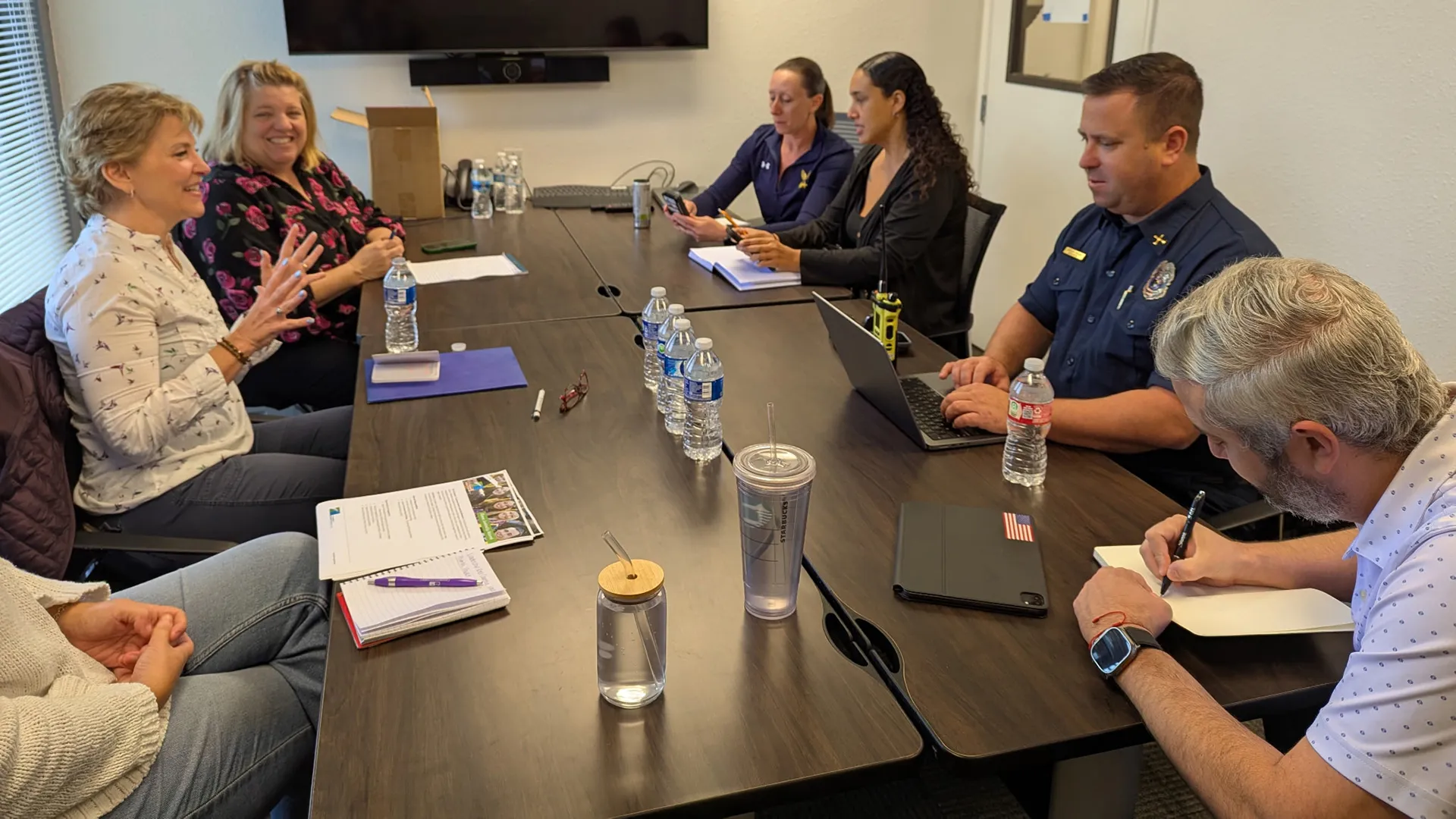 Mona speaking to a group in a conference room, including a man in uniform with a walkie talkie